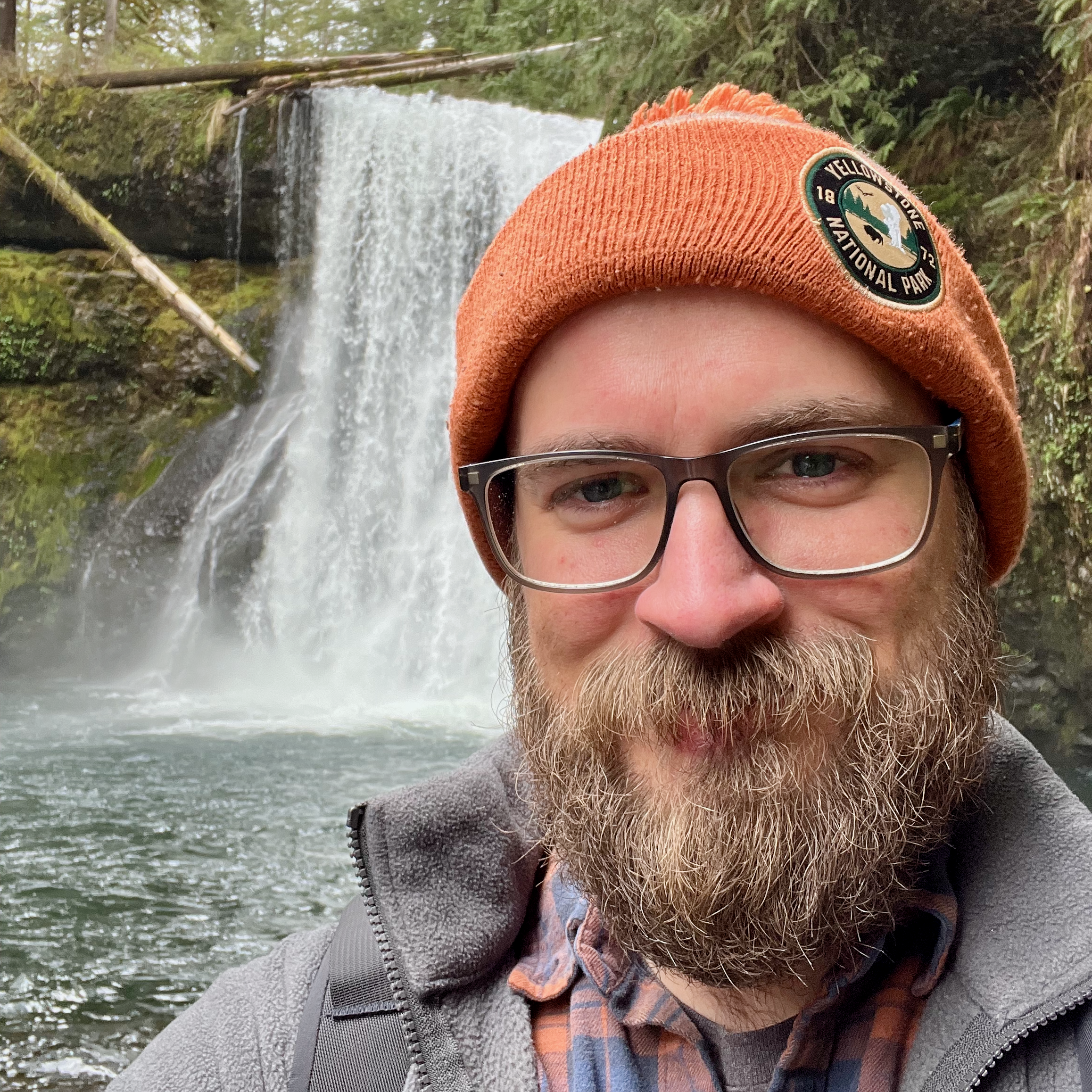 Portrait of a man with a beard, wearing a hat, standing in front of a waterfall.